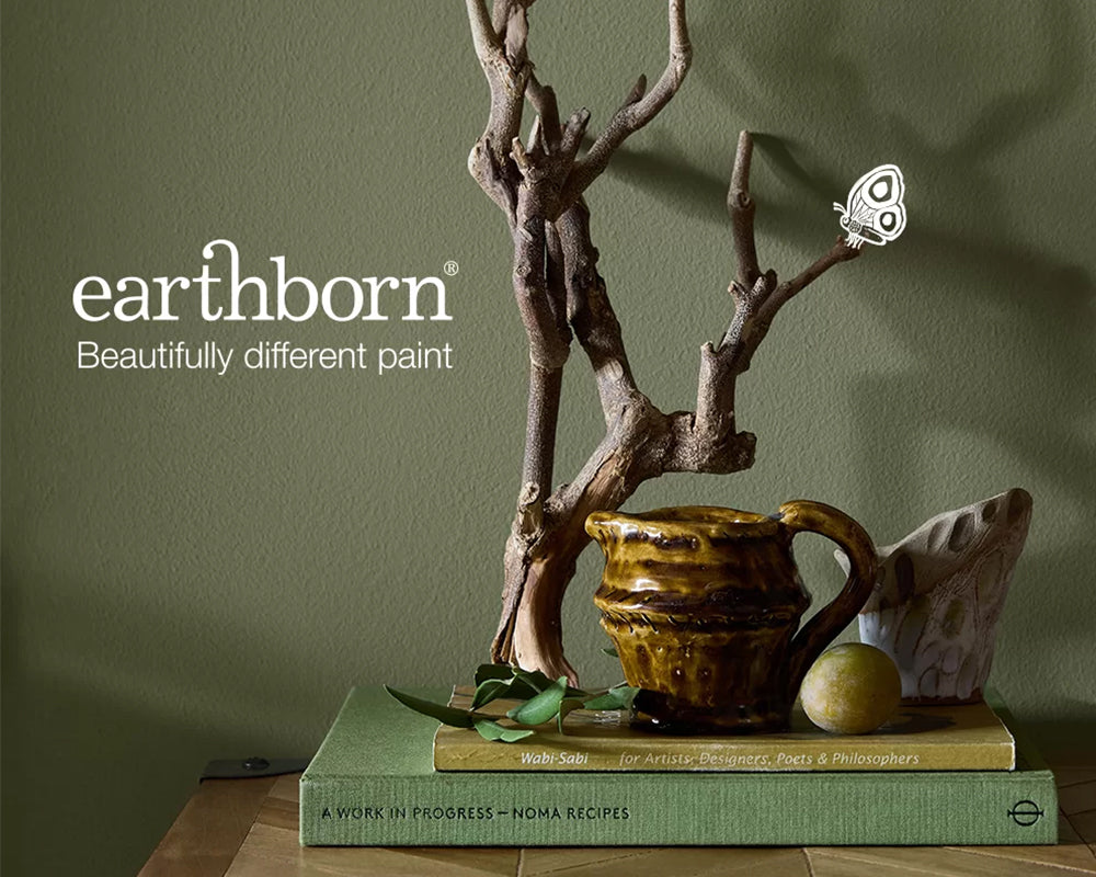 Decorative setup with a jug, books, and branches on a table against a green wall of the Oliver Twig colour with 'earthborn' branding.