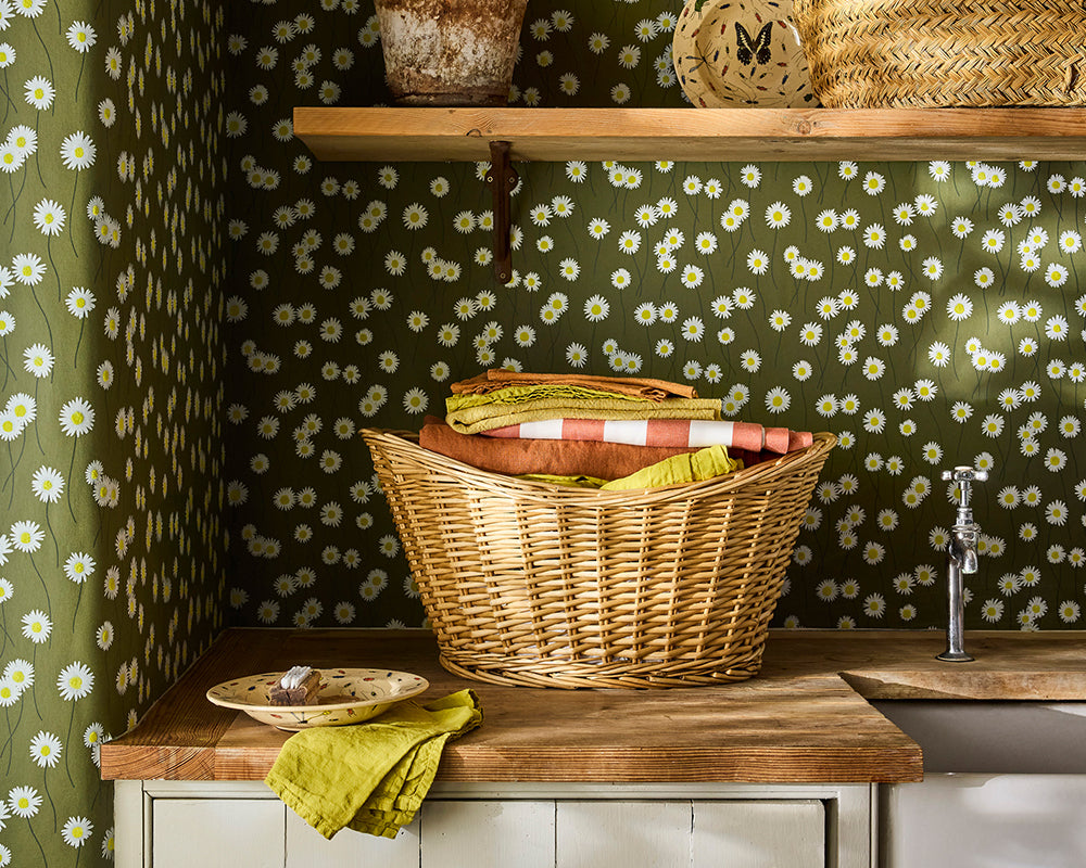 Wicker basket with folded clothes on a wooden surface against a floral wallpapered wall, Little Greene Moon Daisies Wallpaper