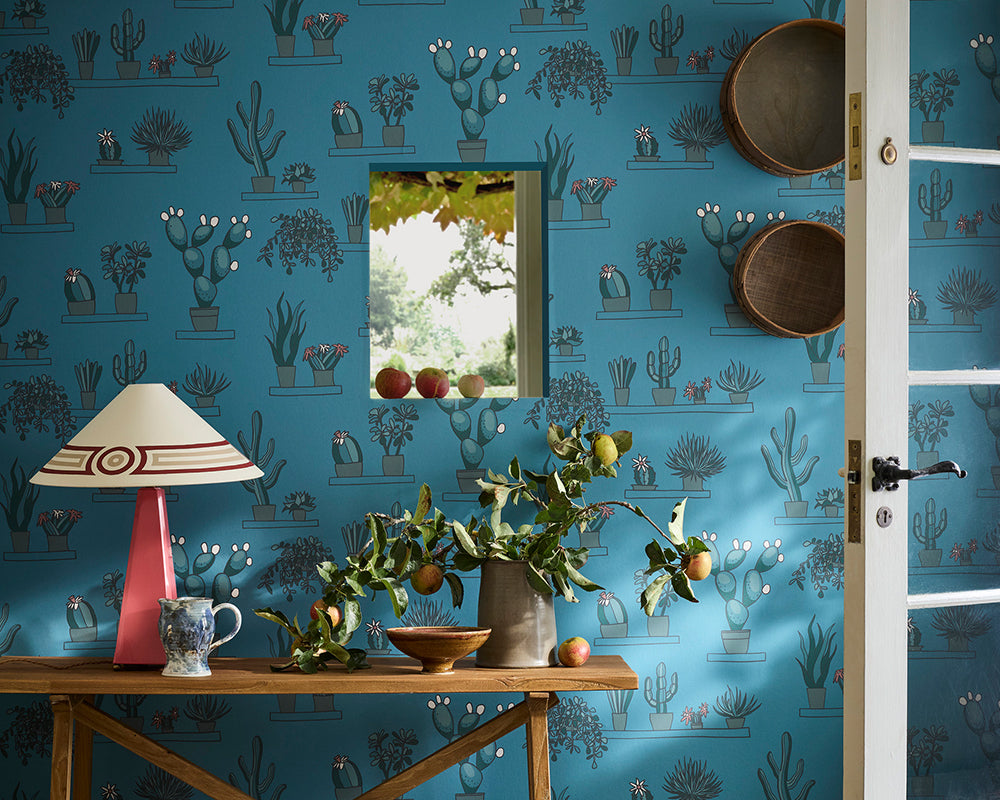 Room interior with blue cactus-patterned Little Greene Mr Straw's Greenhouse Wallpaper, wooden table, lamp, and plants.