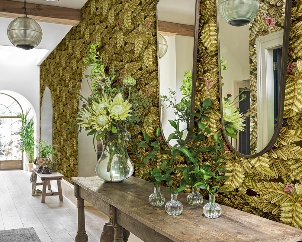 Decorative interior with floral arrangements, mirrors, and a wooden table against the leaf-patterned Cole & Son Acacia Canopy Wallpaper.