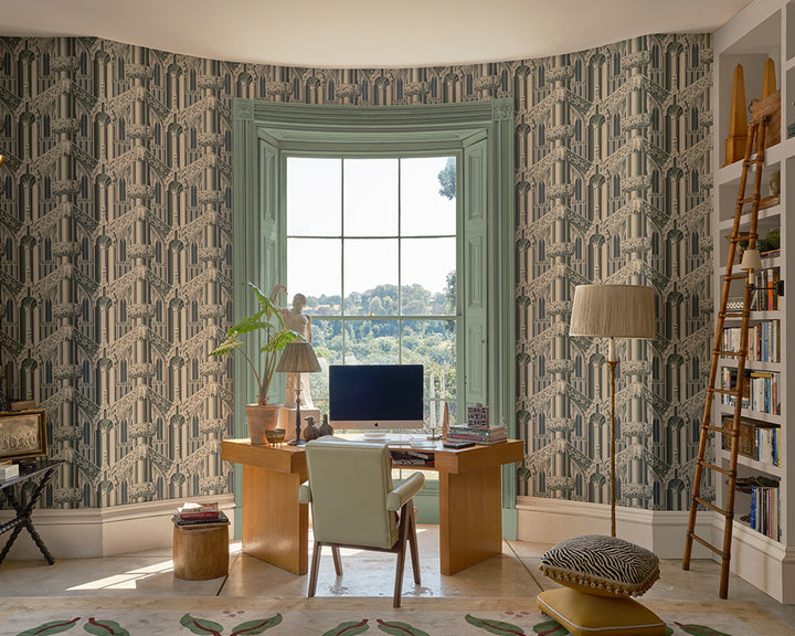 A styled home office featuring Farrow & Ball Gothic wallpaper with a repeating geometric art deco pattern in muted green, beige, and gray tones. The room includes a wooden desk, a light green chair, a window looking out onto a garden, a wooden ladder shelf, and a patterned pouf.