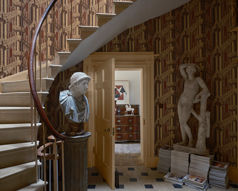 An ornate interior staircase with a curved wooden handrail, patterned Farrow & Ball Gothic wallpaper, a white marble bust sculpture, a full-length white marble statue, and stacks of magazines on the floor.