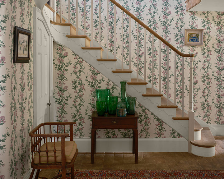 A classic interior staircase with white balusters and wooden steps, set against Sweet Pea, floral wallpaper. A small wooden console table under the stairs holds green glass vases, next to a wooden spindle-back chair with a beige cushion.