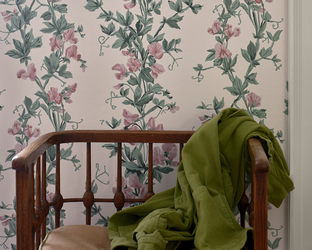 Sweet Pea floral wallpaper with pink flowers and green leaves on a light background, shown behind a wooden chair with a green jacket draped over it.