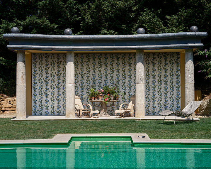 A stone patio with Sweet Pea, blue and white floral wallpaper, columns, a small table with potted plants, and two chairs, situated behind a swimming pool with a lawn and trees in the background. 