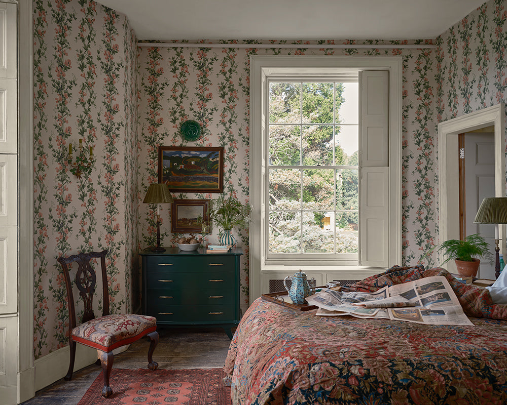 A vintage-style bedroom with the floral  Sweet Pea wallpaper, a dark green chest of drawers, a wooden chair, and a bed with a matching floral quilt. A large window in the center shows green trees outside.