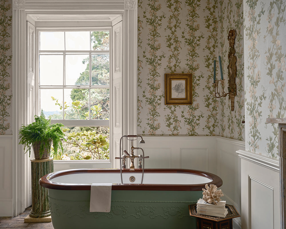 A vintage-style freestanding bathtub in a beautifully decorated bathroom with the floral wallpaper, Sweet Pea, from Farrow & Ball, white wainscoting, a large window, and a potted fern.