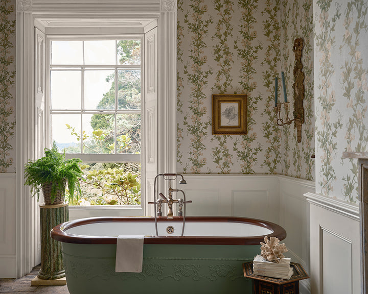 A vintage-style freestanding bathtub in a beautifully decorated bathroom with the floral wallpaper, Sweet Pea, from Farrow & Ball, white wainscoting, a large window, and a potted fern.