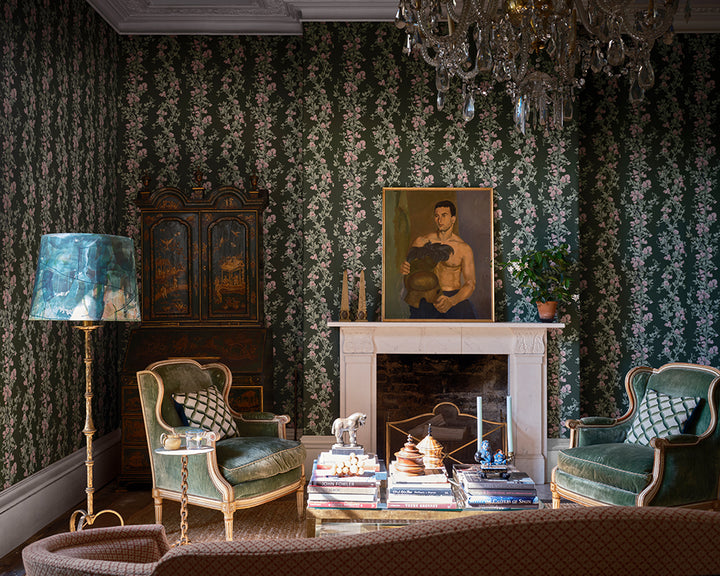 An elegant vintage living room featuring dark green floral wallpaper, Sweet Pea from Farrow & Ball, two green velvet armchairs with patterned pillows, a white fireplace mantel with a framed portrait, a gold cabinet, and a crystal chandelier.