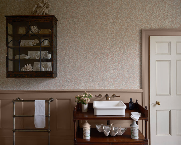 A vintage-style bathroom interior featuring floral wallpaper, Farrow and Ball Vine Leaf, wainscoting, a dark wood wall cabinet, a white pedestal sink on a wooden cart, and a white paneled door.