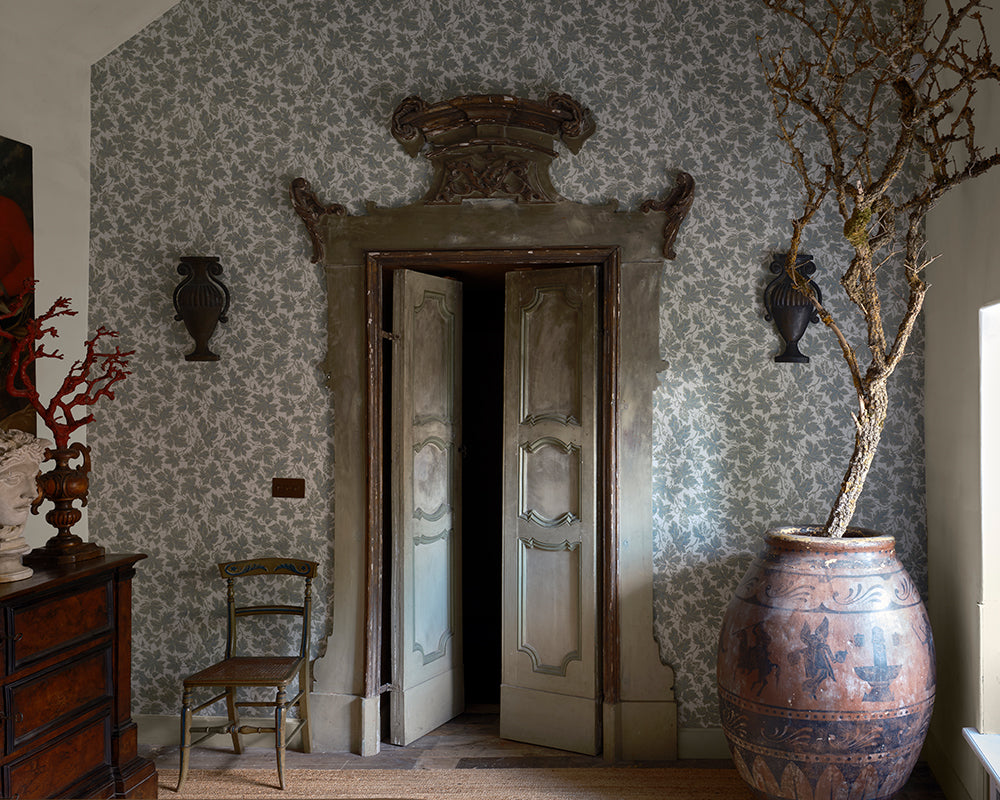 An ornate interior scene with patterned wallpaper, Vine Leaf from Farrow & Ball, a large carved stone doorway opening to a dark room, a wooden chair, wall-mounted vases, and a large terracotta planter holding a bare tree trunk.