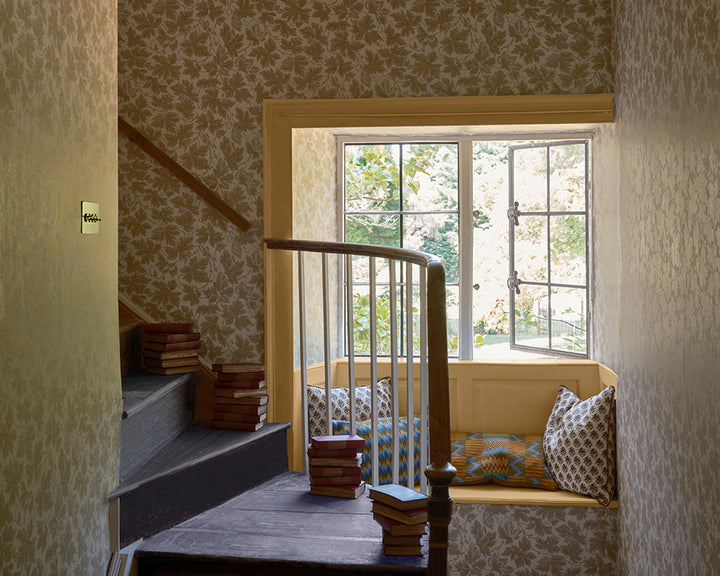An interior room scene with patterned wallpaper, Farrow and Ball Vine Leaf, a wooden staircase, a window seat with pillows, and several stacked books on the steps and bench. 