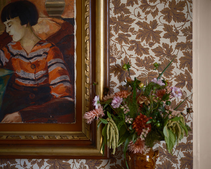 A close-up of a framed painting of a woman in an orange and black striped sweater, set against brown and beige botanical wallpaper, Vine Leaf by Faroow & Ball, with a vase of flowers in the foreground.