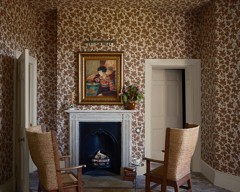 A classic interior room featuring Farrow and Ball Vine Leaf floral patterned wallpaper, a white fireplace with a gold-framed painting, two woven wicker armchairs, and white doors.