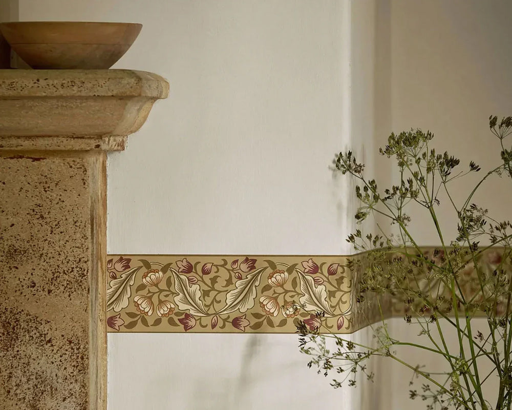 A decorative floral border wallpaper strip with a vintage botanical pattern in muted pink, green, and beige tones, shown on a wall beside a stone mantle and dried greenery.