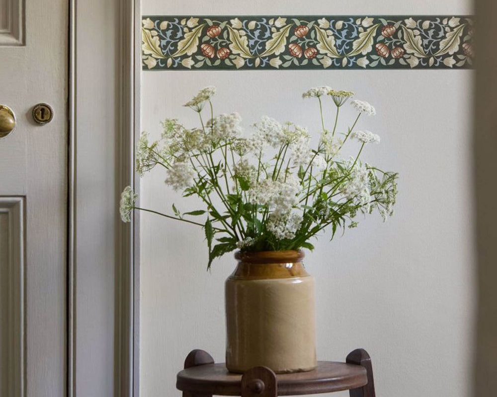 A ceramic vase filled with white flowers and green stems, placed on a small wooden stool against an off-white wall with a decorative border wallpaper strip above.