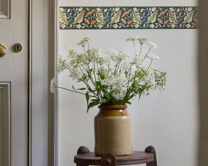 A ceramic vase filled with white flowers and green stems, placed on a small wooden stool against an off-white wall with a decorative border wallpaper strip above.