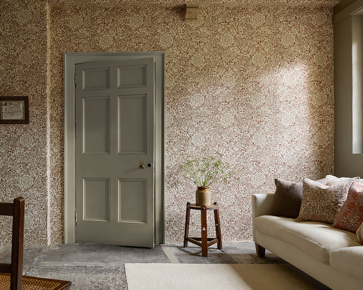 A styled interior room showing a cream sofa with patterned throw pillows, a small wooden side table with a vase of branches, and a wall covered in ornate beige and dusty rose floral wallpaper, Chamomile by Morris & Co. A window and panelled door are visible in the room.