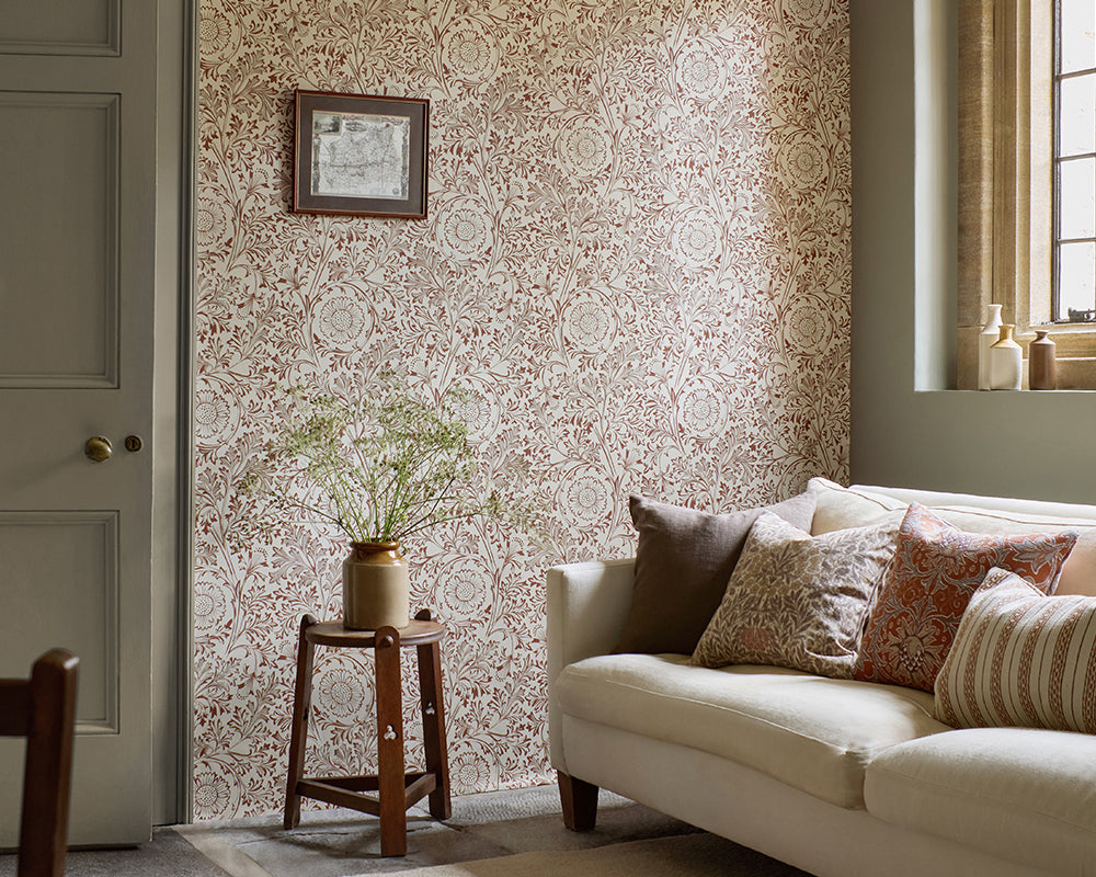 A styled interior room showing a cream sofa with patterned throw pillows, a small wooden side table with a vase of branches, and a wall covered in ornate beige and dusty rose floral wallpaper, Chamomile by Morris & Co. A window and panelled door are visible in the room.