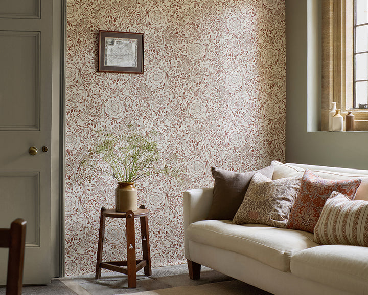 A styled interior room showing a cream sofa with patterned throw pillows, a small wooden side table with a vase of branches, and a wall covered in ornate beige and dusty rose floral wallpaper, Chamomile by Morris & Co. A window and panelled door are visible in the room.