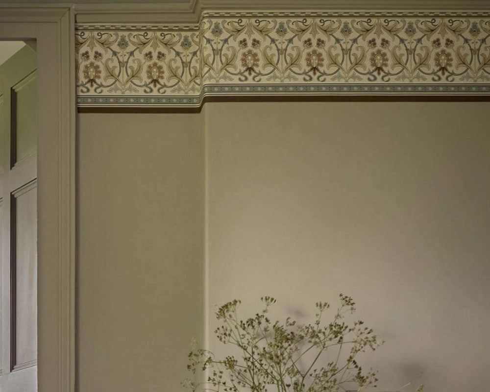 An ornate decorative wallpaper border with a floral and scroll pattern in beige, brown, and muted blue, installed on a wall above a cream-colored door frame.