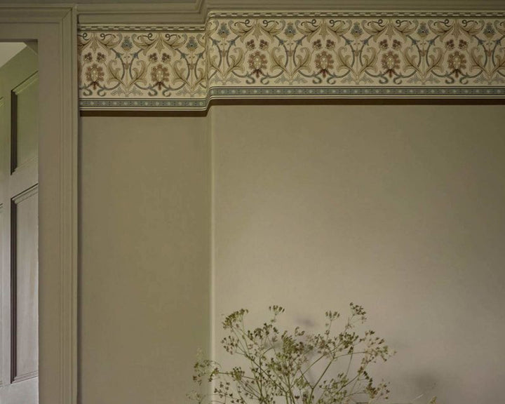 An ornate decorative wallpaper border with a floral and scroll pattern in beige, brown, and muted blue, installed on a wall above a cream-colored door frame.