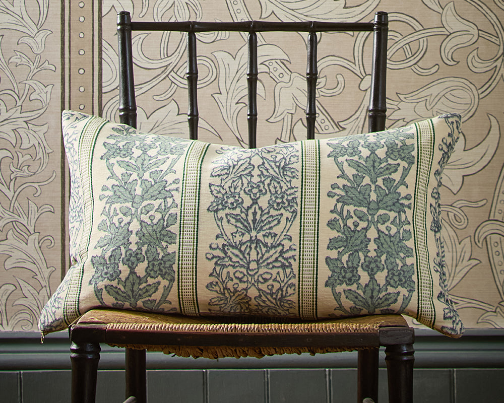 A decorative throw pillow with a blue-green botanical damask pattern on a cream background, displayed on a dark wooden chair against a patterned wallpaper backdrop, Etched Scrolling Tulip by Morris & Co.