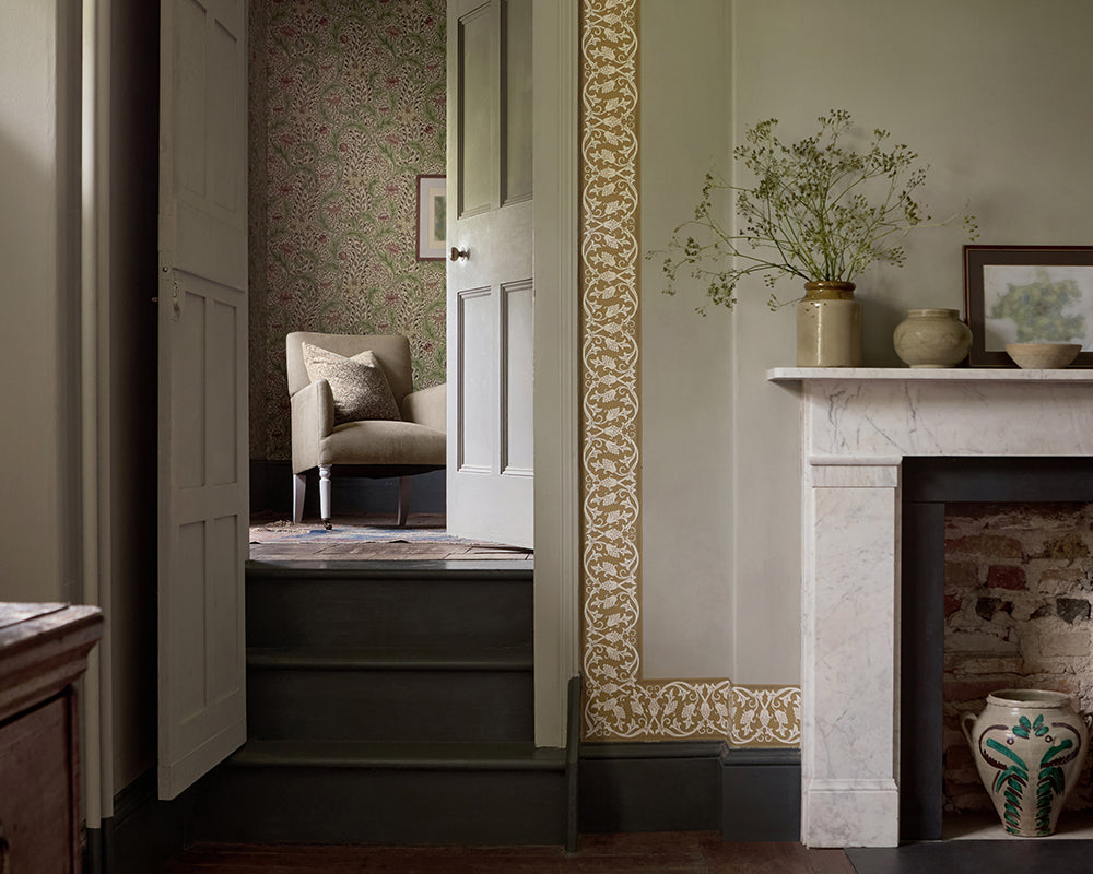 An interior room scene showing a doorway, with St John border wallpapered on the wall, a fireplace mantel with vases and framed art, and a glimpse into an adjacent sitting area with a chair.