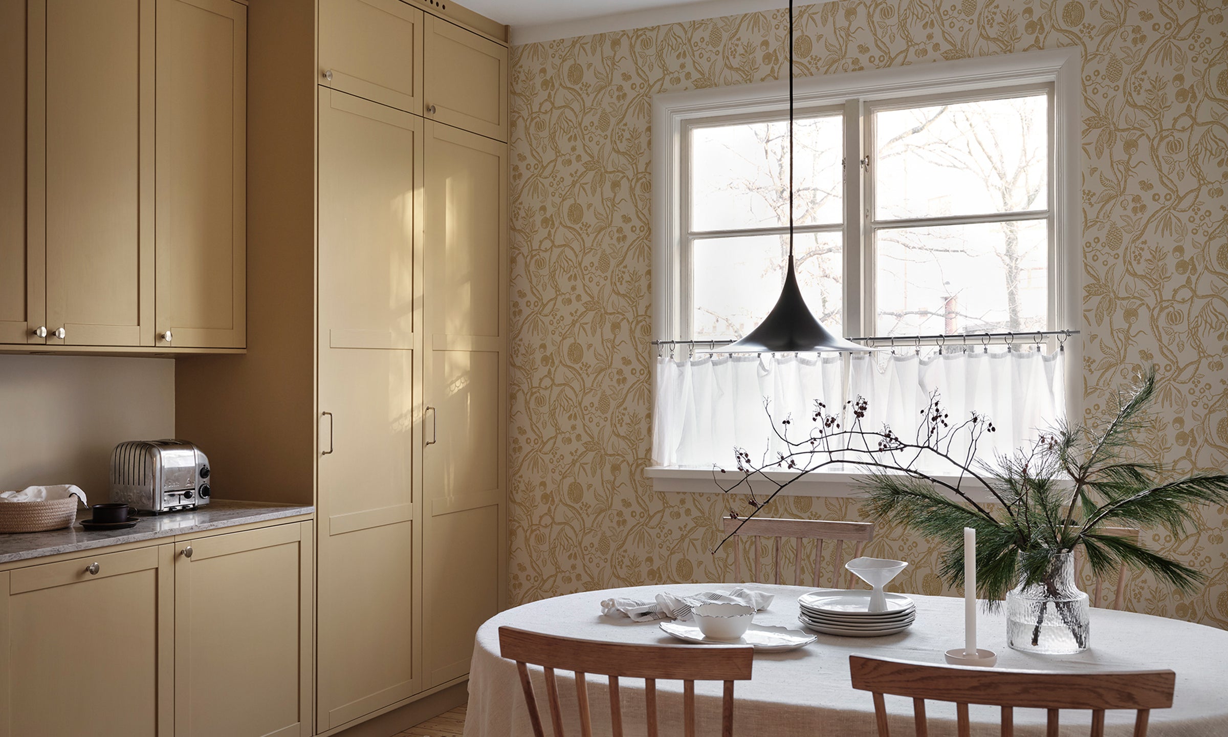 Sandberg Tree of Life Wallpaper in a Kitchen with beige cabinets, a round dining table, and a window with a decorative curtain.