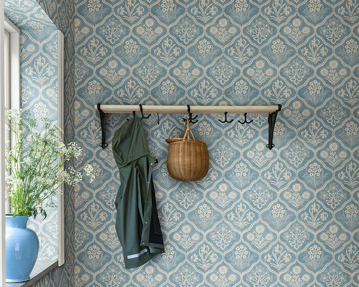 patterned wallpaper wall with hooks with a green coat and basket, and a plant.